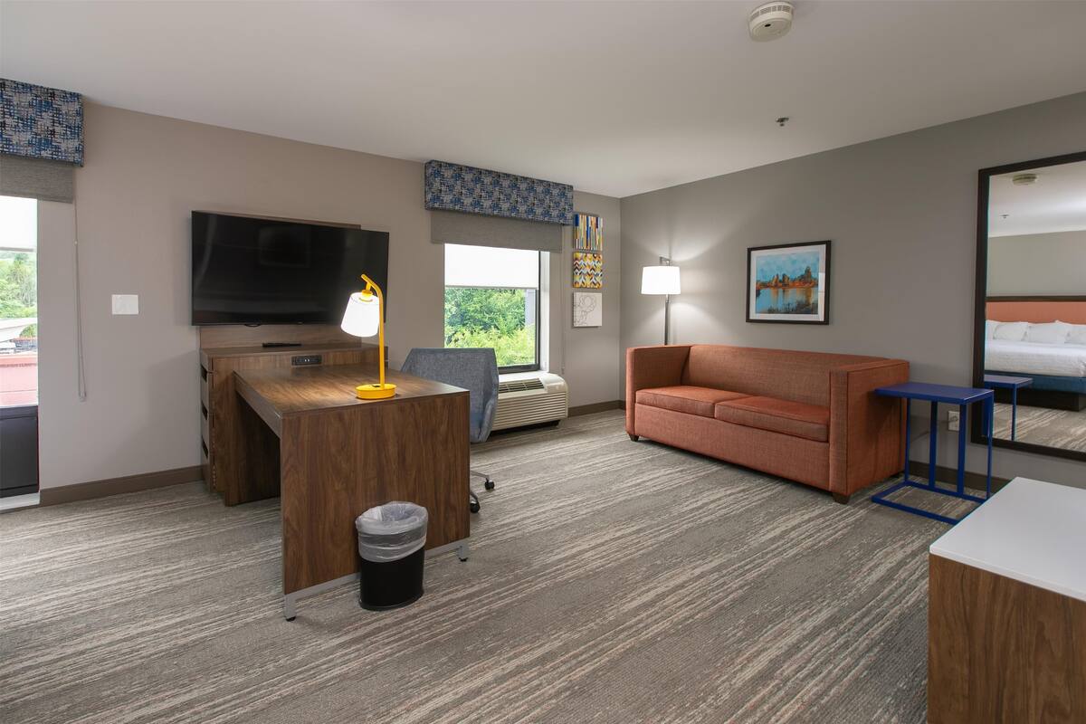 A hotel room with a wooden desk, a flat-screen TV, a sofa, a lamp, a painting, and a bed with white linens.