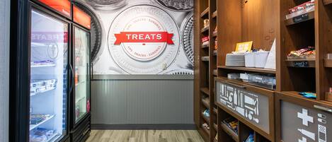 A store interior with a 'Treats' sign, refrigerated display cases, and wooden shelves stocked with snacks and sundries.