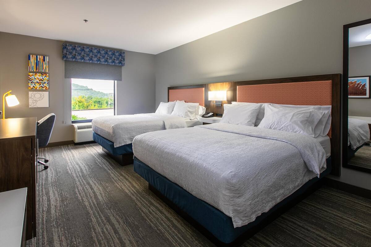 A hotel room with two beds, a desk, a chair, a window with a view, and artwork on the walls.