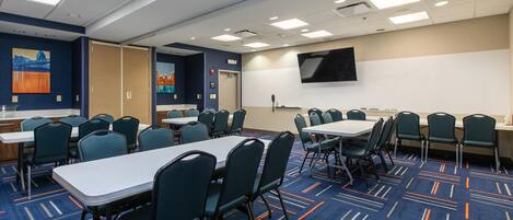 A conference room with multiple tables and chairs, a flat-screen TV mounted on the wall, and a carpeted floor.