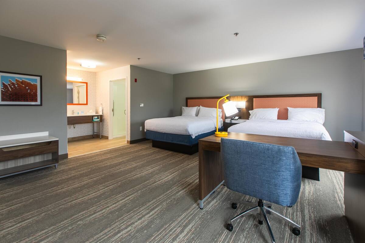 A hotel room with two beds, a desk, a chair, a mirror, and a bathroom visible in the background.