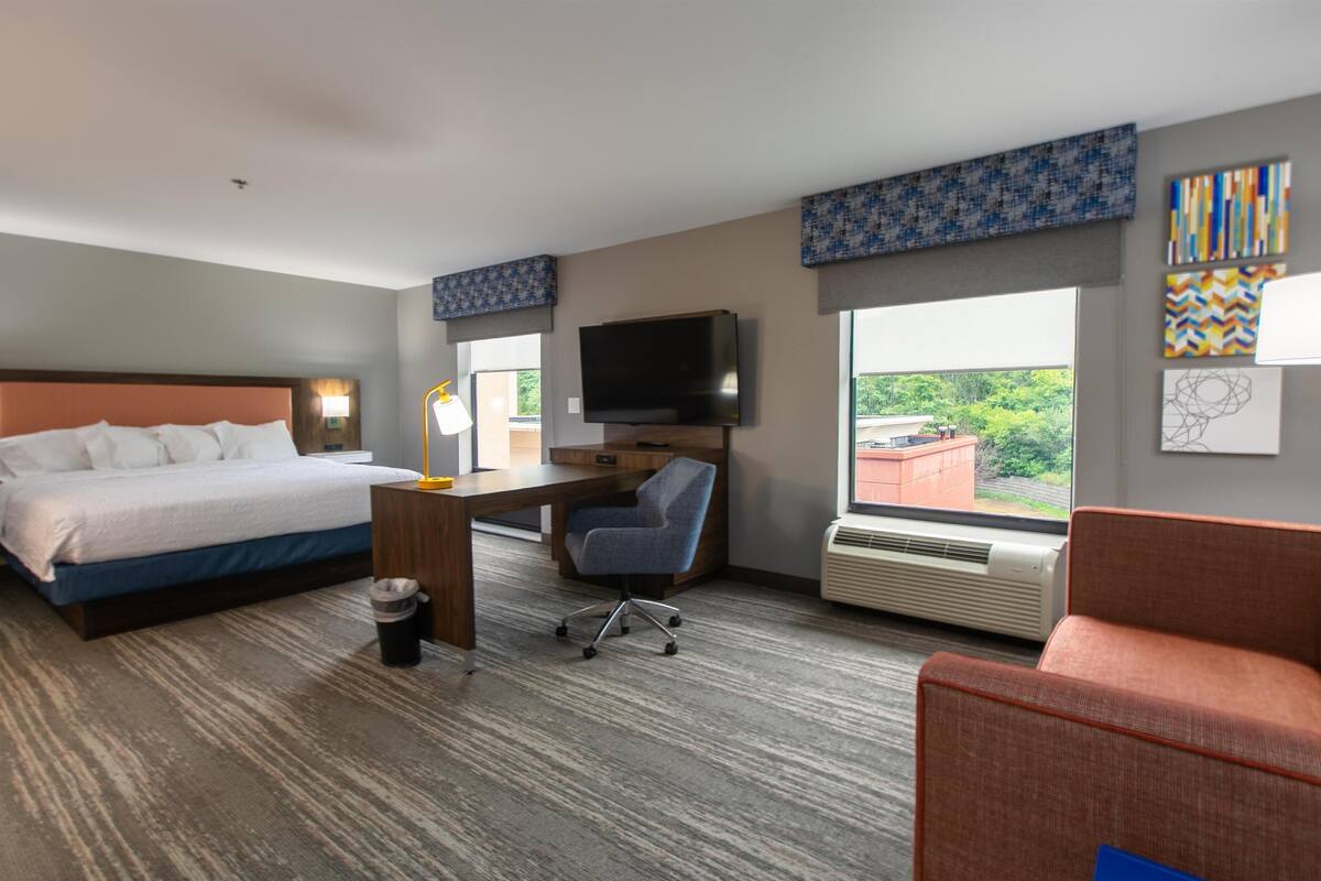 A hotel room with a bed, desk, chair, TV, and large windows.