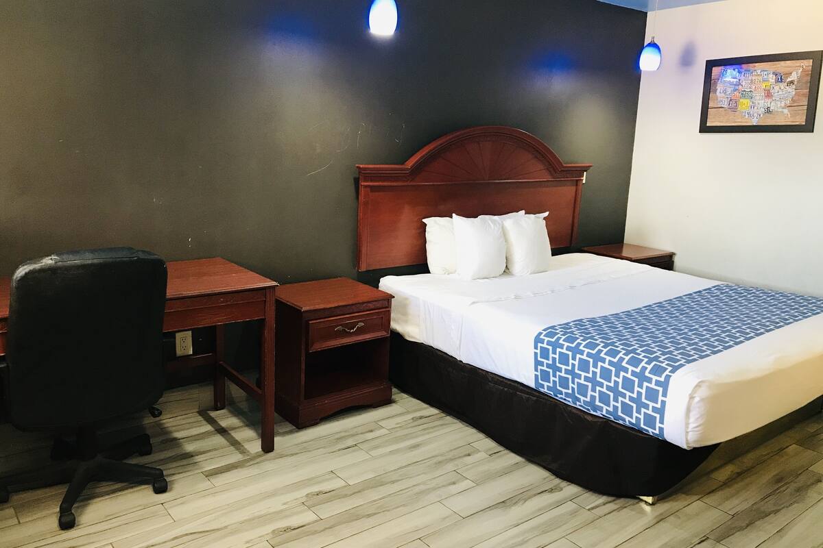 Quarto de hotel com cabeceira de madeira, cama com roupa de cama branca e manta azul estampada, mesa de cabeceira de madeira, cadeira de escritório preta e duas luminárias pendentes.