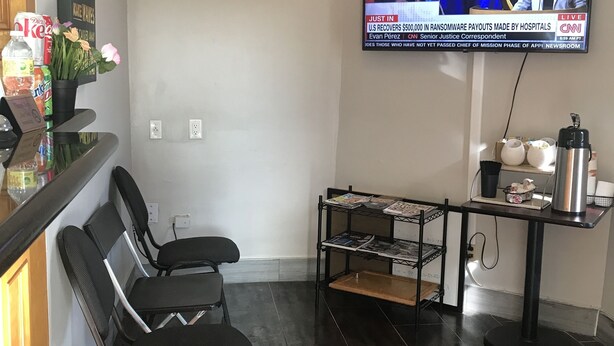 A modern living room with a flat-screen TV displaying a news broadcast, a coffee table, a bookshelf, and a seating area with chairs. - Summit Inn - Houston Medical Center (Houston)