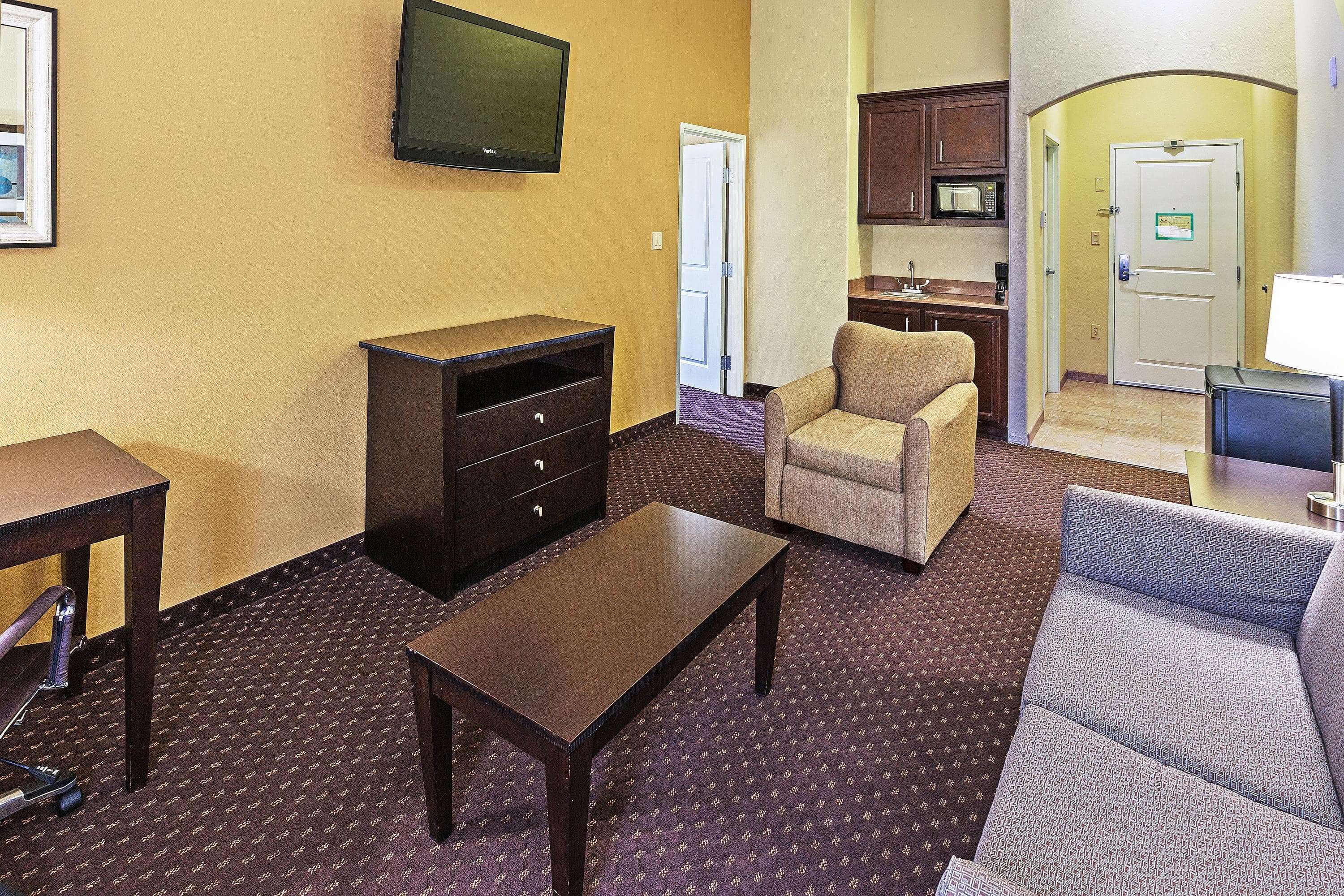 A hotel room with a television, a sofa, a chair, a desk, and a kitchenette.