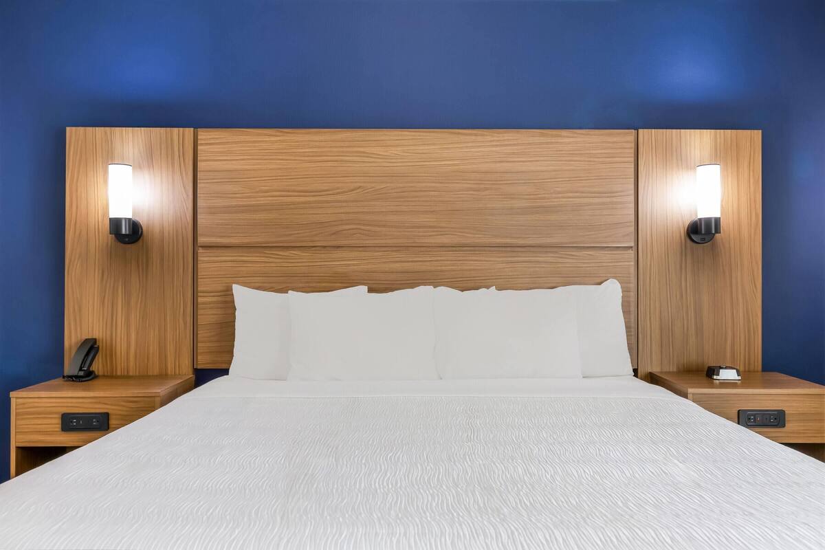 Ein Hotelzimmer mit einem hölzernen Kopfteil, zwei Nachttischen mit Lampen, einem Bett mit weißer Bettwäsche und einer Bedienkonsole am Bett.