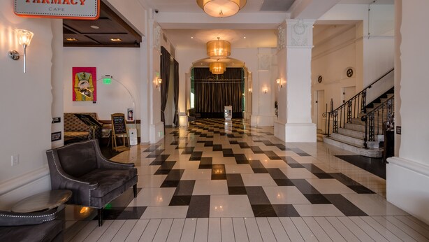 Un vestíbulo de hotel con suelo a cuadros, una mesa de recepción, un rótulo de farmacia y una escalera.
