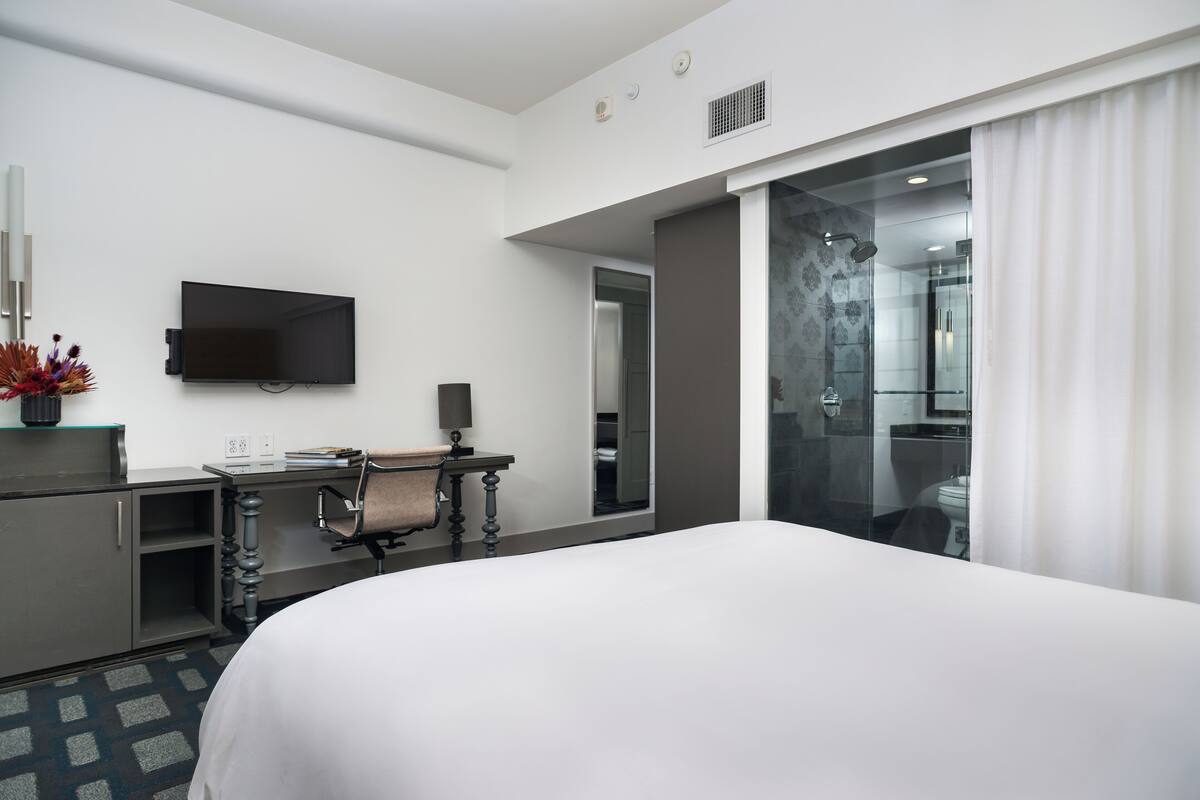 A hotel room with a large bed, a desk with a chair, a flat-screen TV, and a shower area.