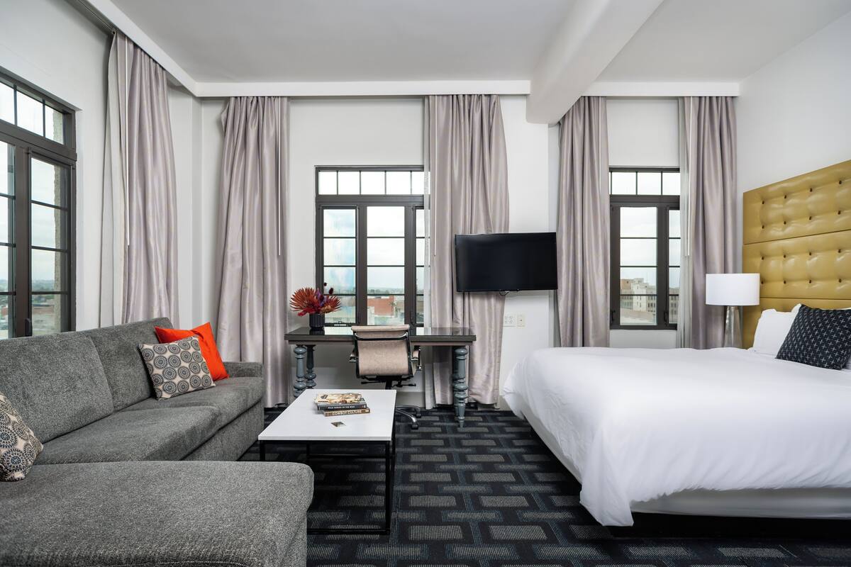 A hotel room with a bed, a sofa, a desk with a chair, a TV, and large windows with curtains.