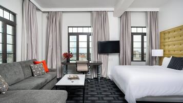 Habitación de hotel con cama, sofá, escritorio con silla, televisor y ventanas grandes con cortinas.