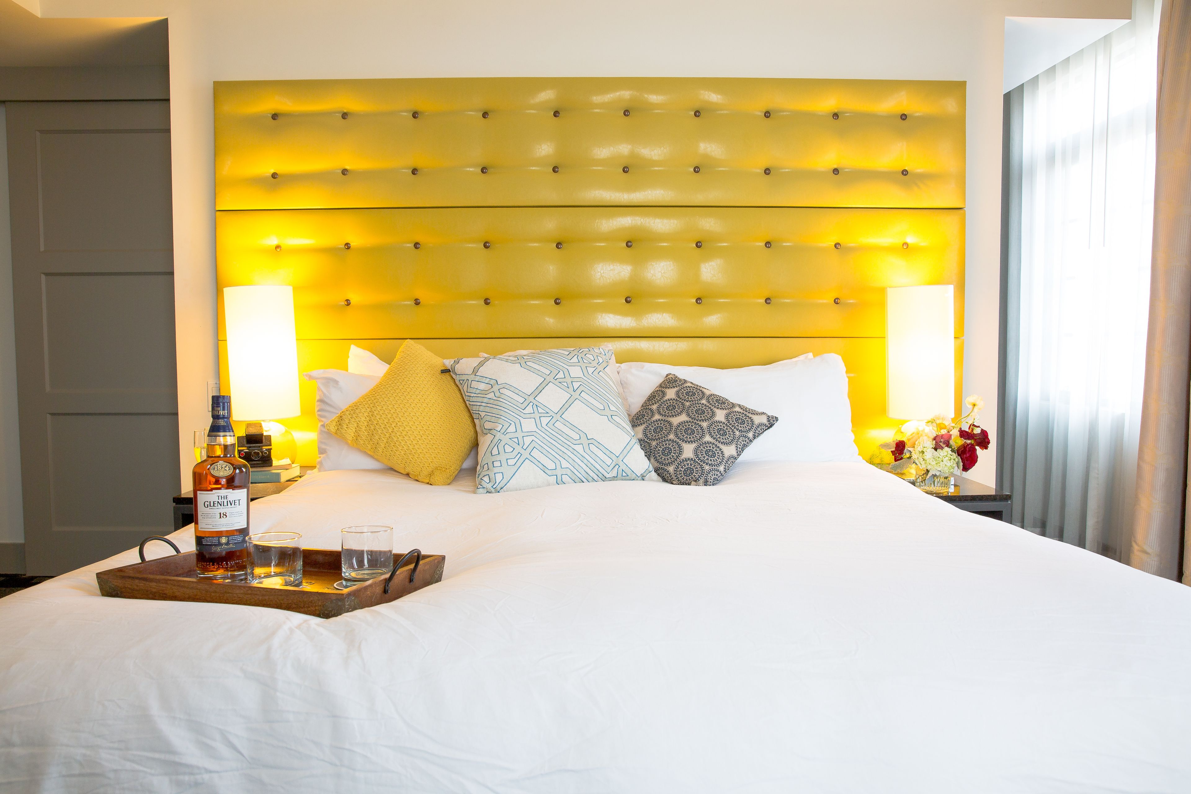 A hotel room with a large bed, a headboard with a yellow panel, a tray with a bottle and glasses, and a vase with flowers.