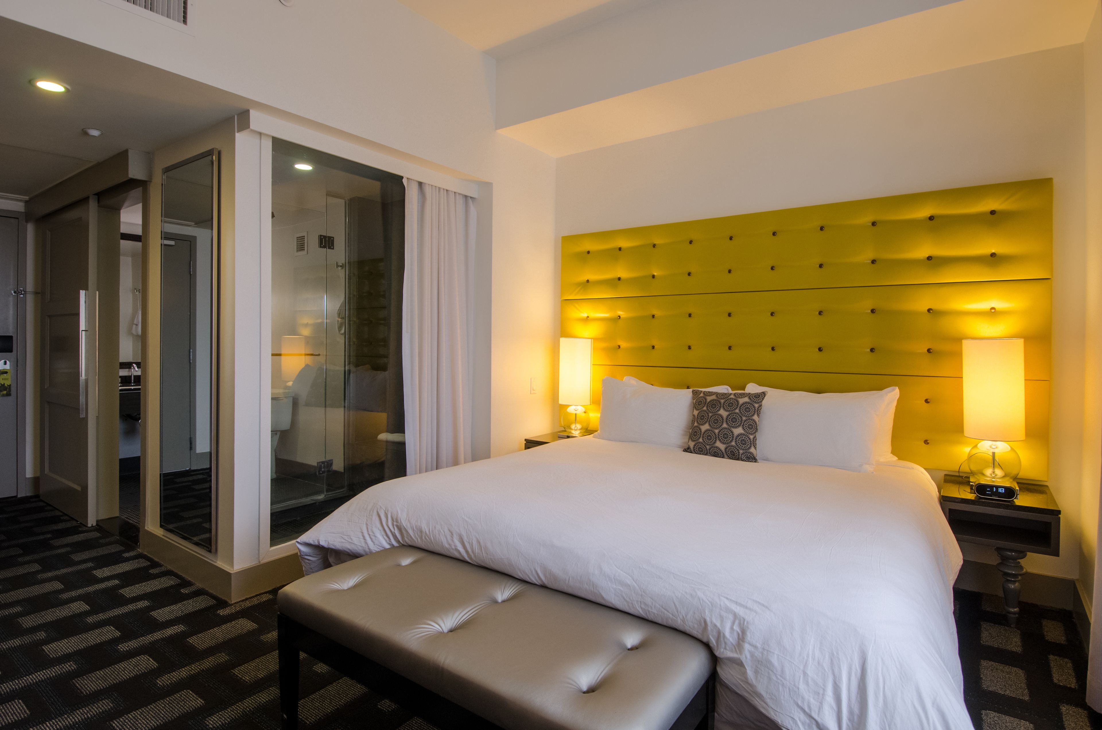 A hotel room with a large bed, a bench, a glass door leading to a bathroom, and a yellow headboard.