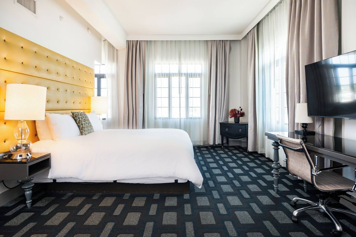 A hotel room with a large bed, a desk with a chair, a television, and a window with curtains.