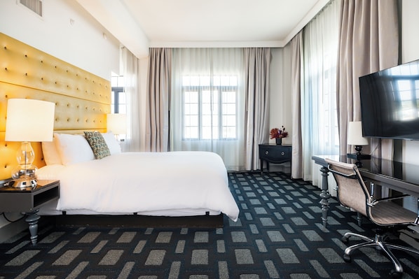 A hotel room with a large bed, a desk with a chair, a television, and a window with curtains. - The Padre Hotel (Bakersfield)