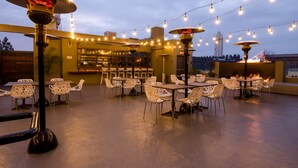 Un patio al aire libre con mesas, sillas, luces colgantes y una zona de bar al fondo.