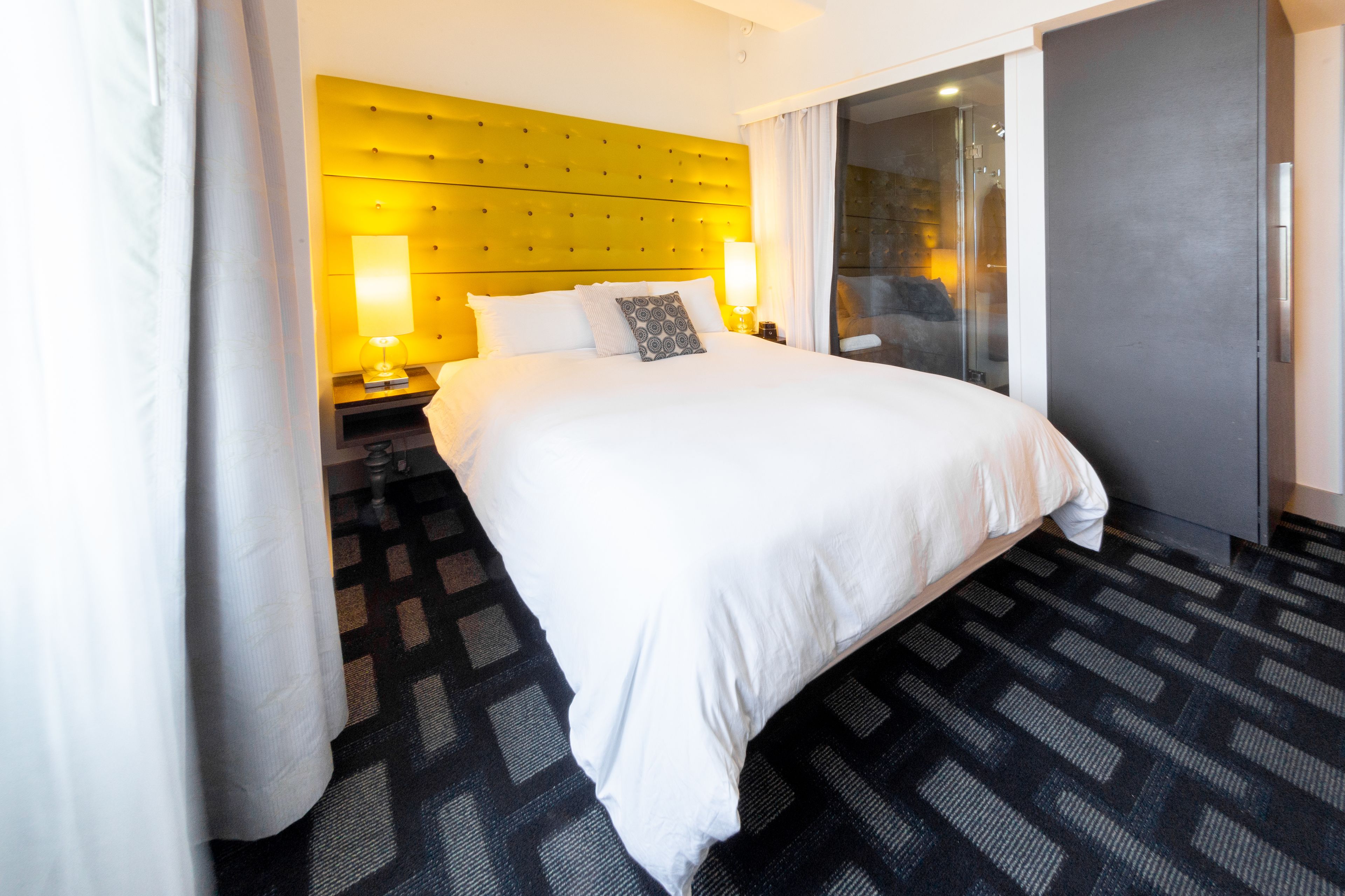 A hotel room with a large bed, a yellow headboard, a glass door leading to a balcony, and a patterned carpet.