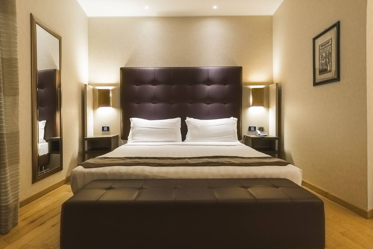 Una habitación de hotel moderna con una cama grande, dos mesitas de noche, un tocador, un espejo amplio y un cuadro en la pared.