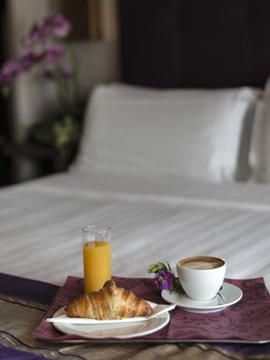 A neatly made bed with white linens, a glass of orange juice, a croissant, and a cup of coffee on a table with a purple napkin.
