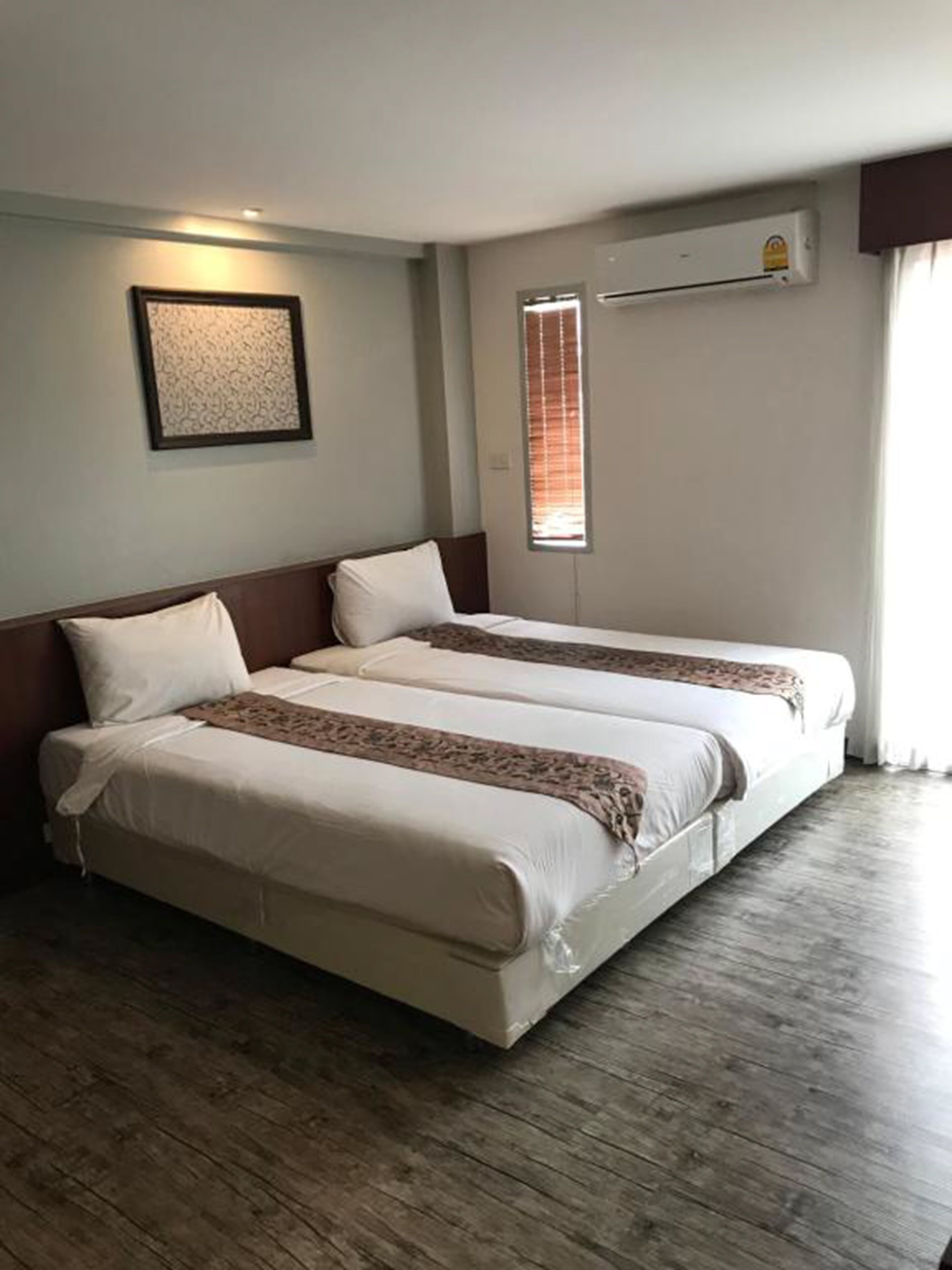 A hotel room with two beds, a framed artwork on the wall, an air conditioning unit, and a window with blinds.