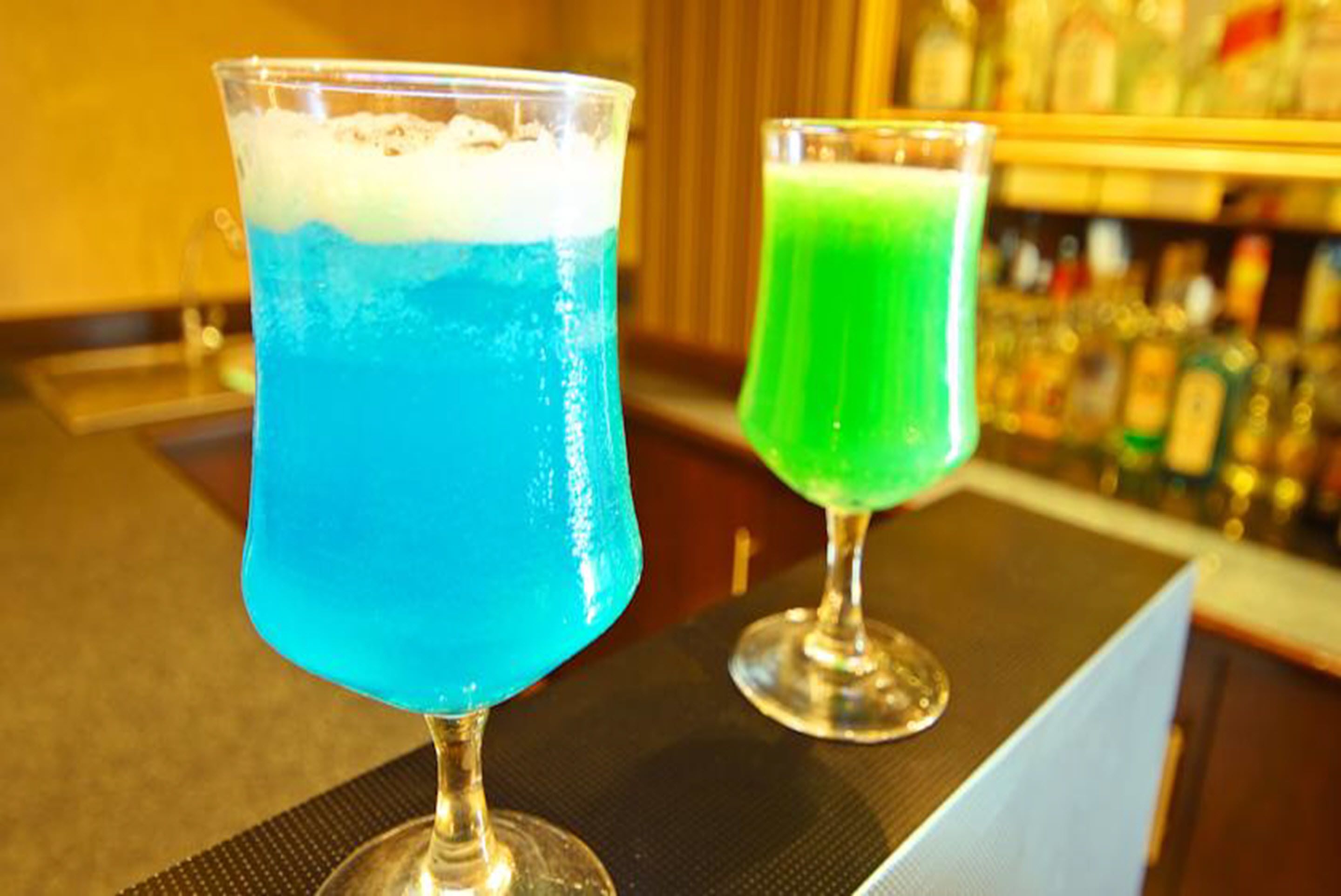 To cocktails i glas på en bardisk med en uskarpt baggrund af flasker.