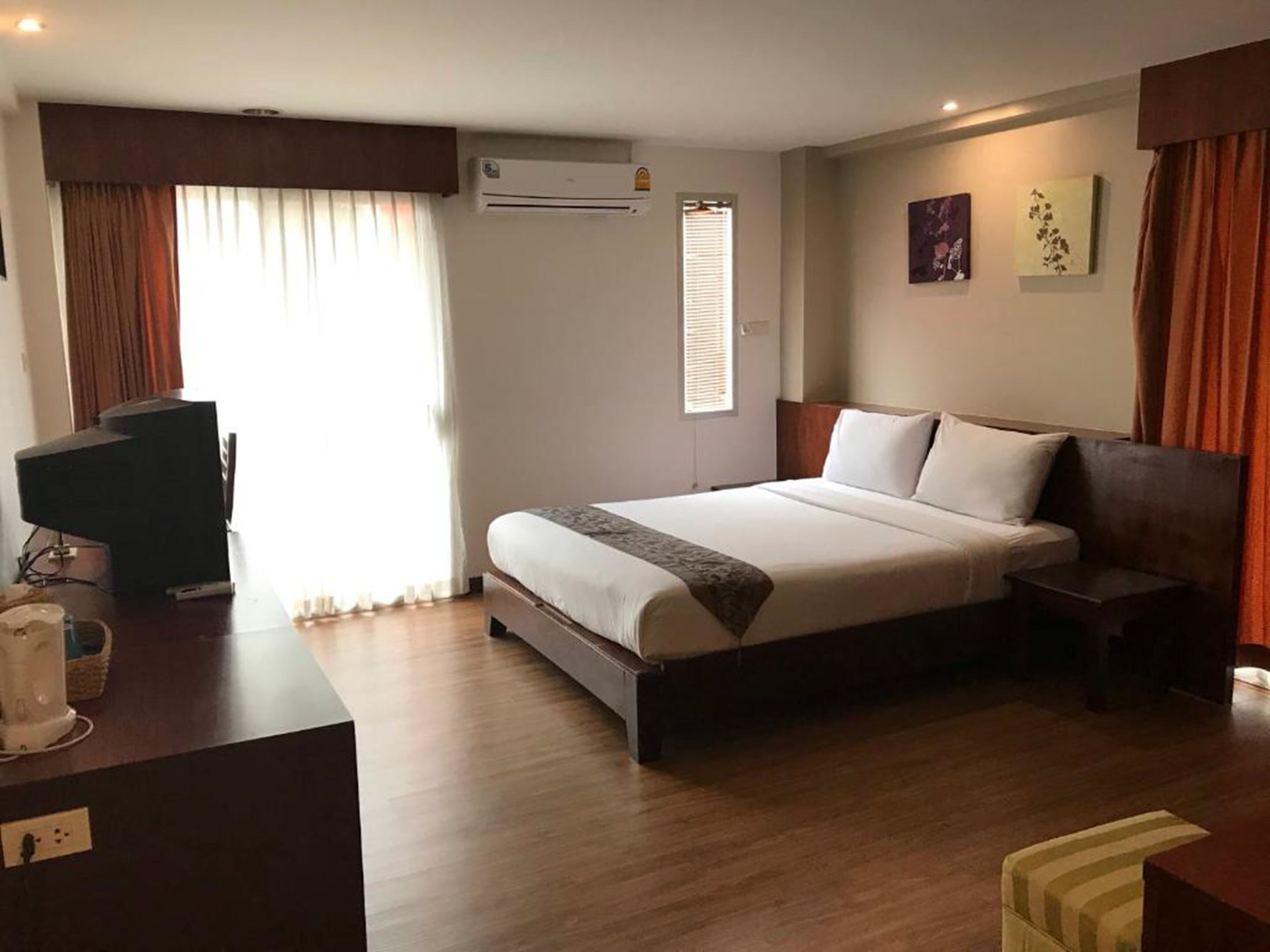 A hotel room with a bed, bedside tables, a desk with a computer, an air conditioner, and a window with curtains.