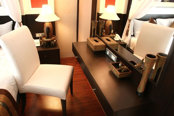 A hotel room with a wooden floor, a white upholstered chair, a bedside table with a lamp, a mirror, and a small wooden box.