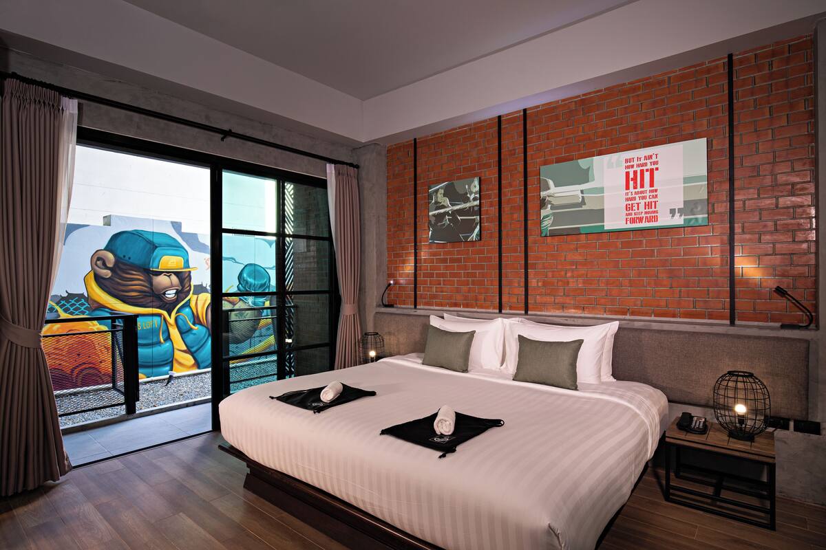 A hotel room with a large bed, a view of a cityscape, a brick wall, and artwork on the wall.