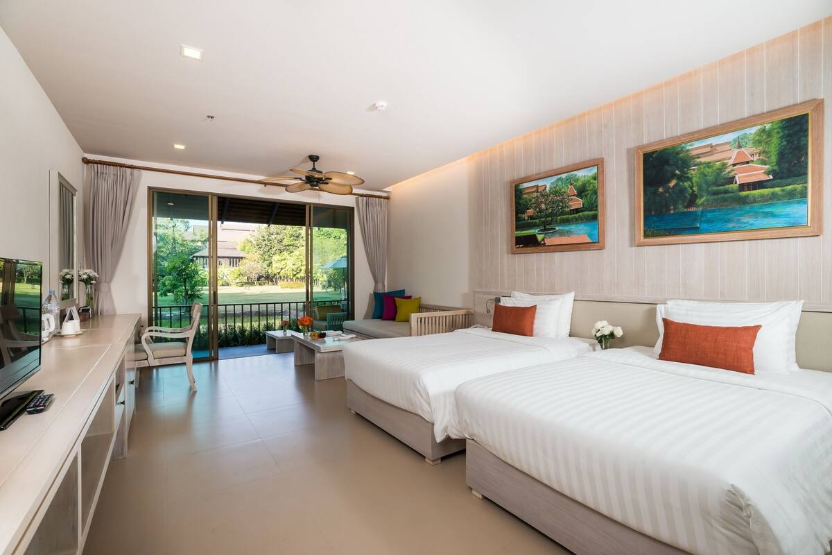 A hotel room with two beds, a desk, a chair, and a balcony with a view of greenery.