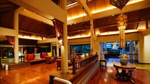 Un hall espacioso con suelo de madera, una zona de estar central con sofás y sillones, un pilar decorativo y vistas al exterior.