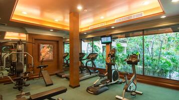 Un gimnasio con diverso equipo de ejercicio, amplias ventanas con vista a zonas verdes y una pantalla digital que muestra los números de las habitaciones.