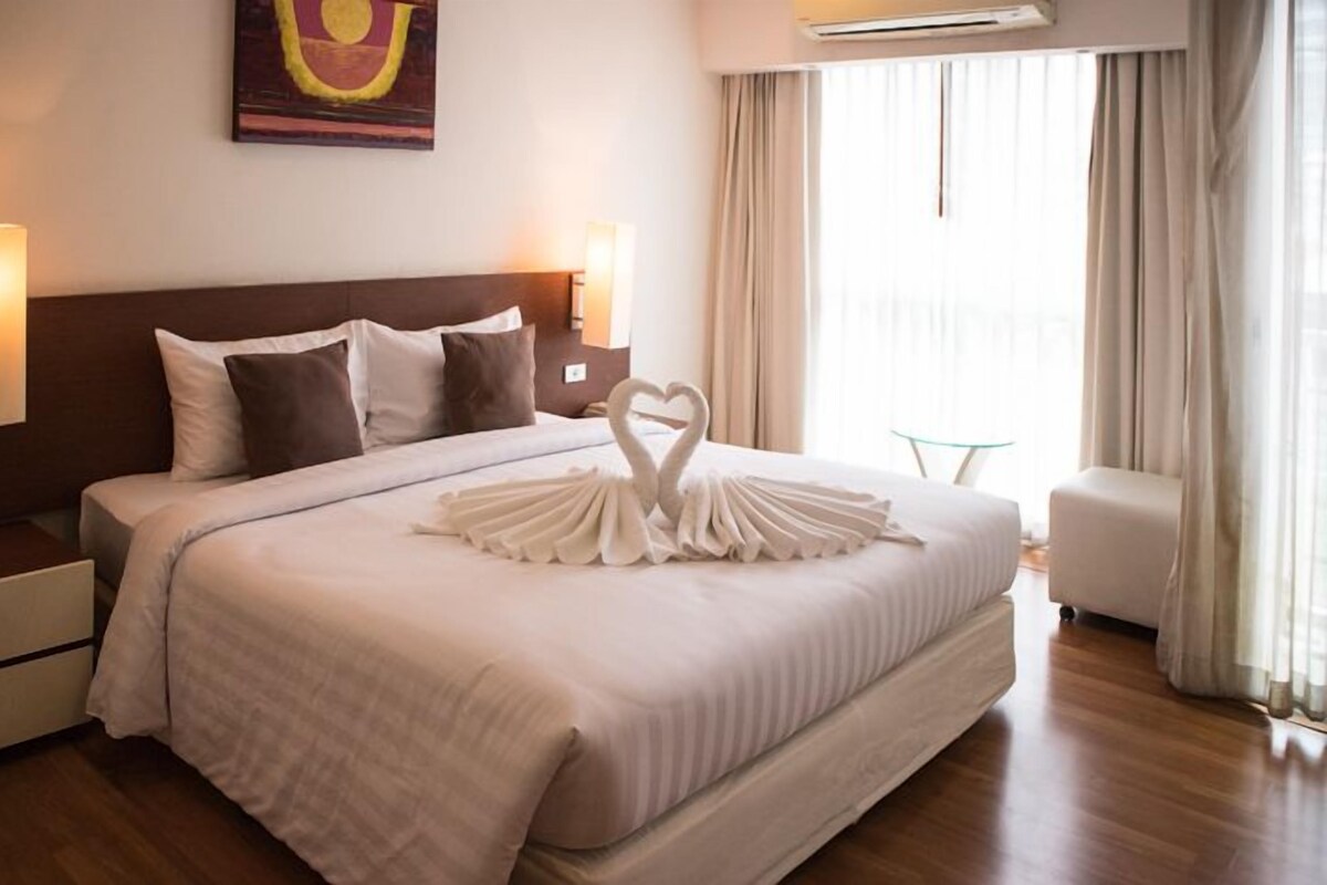 A hotel room with a large bed, two bedside tables, a wall-mounted air conditioner, and a window with curtains.