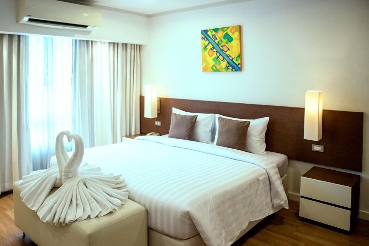A hotel room with a large bed, a bedside table, a nightstand, a wall-mounted lamp, and a decorative towel arrangement.