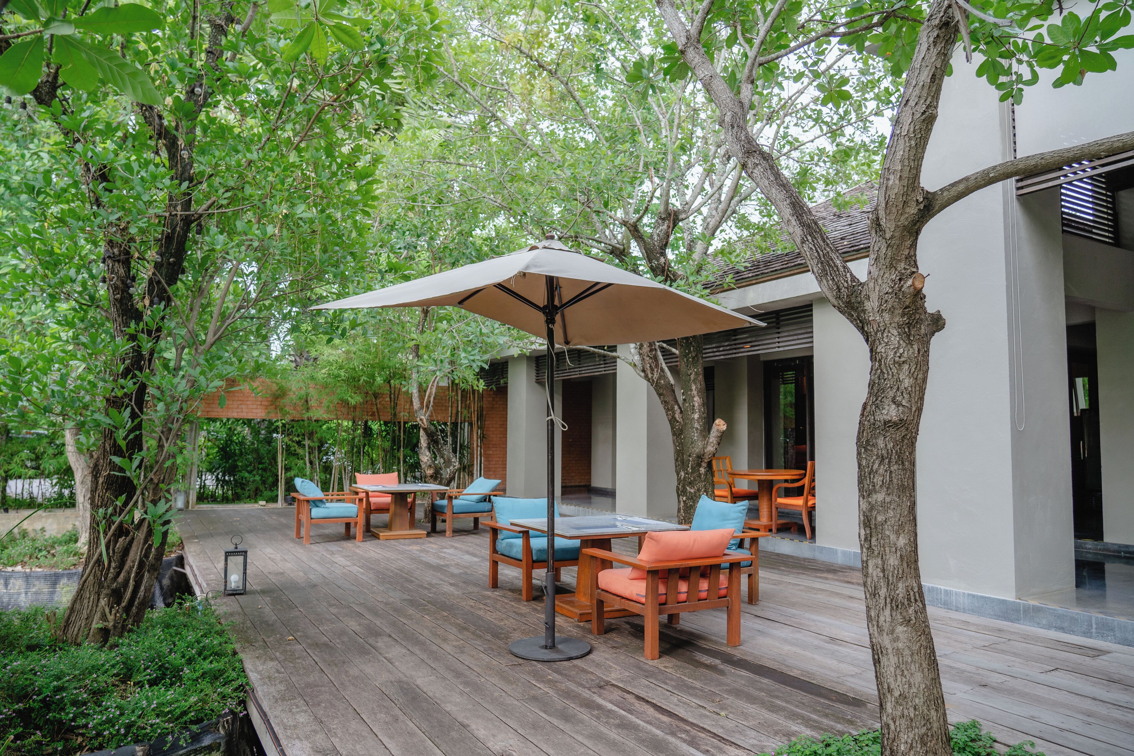 Un patio al aire libre con mobiliario de madera, incluyendo mesas y sillas, y un paraguas grande que ofrece sombra.