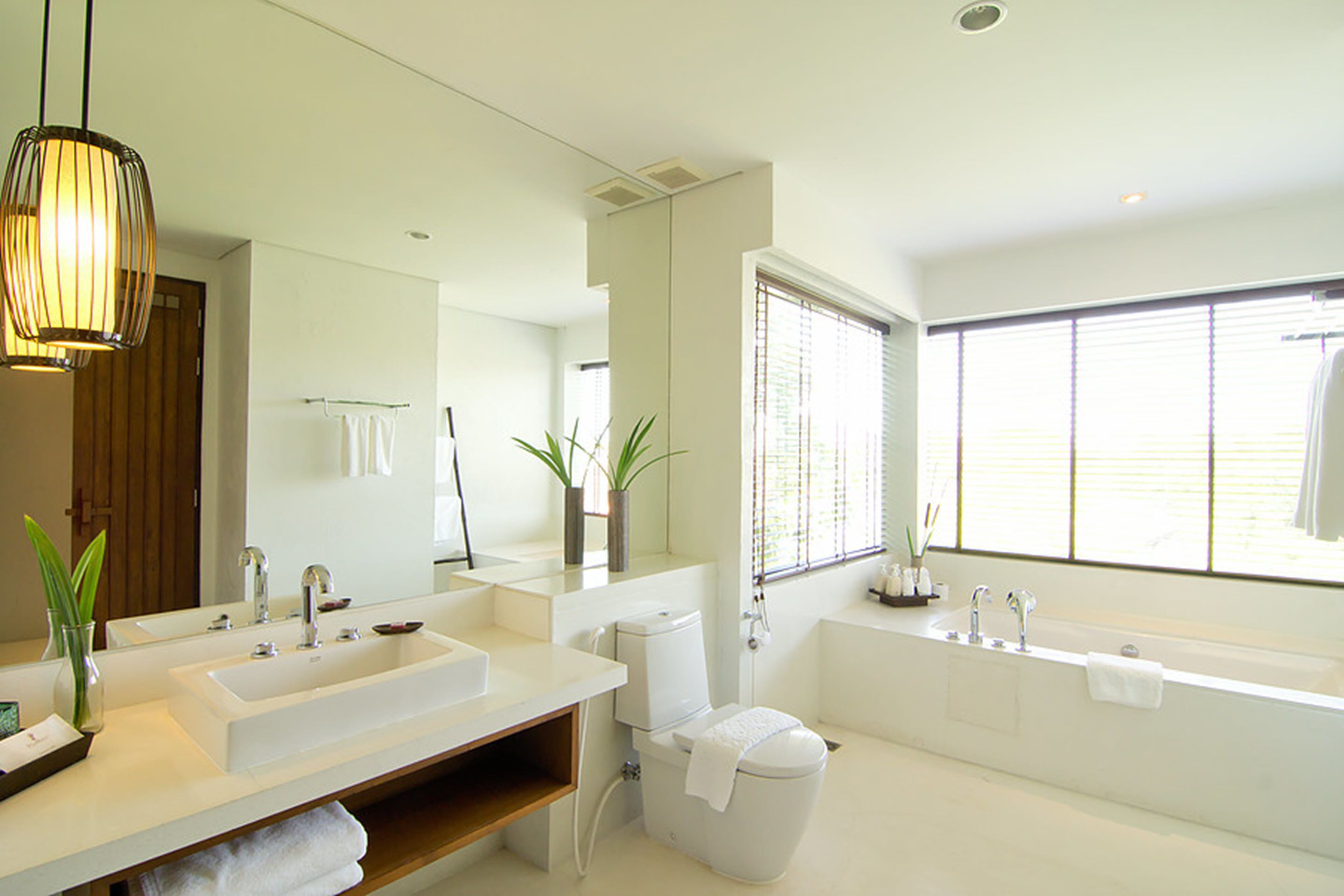 Une salle de bain moderne avec un double vasque, des toilettes et une baignoire. Des plantes sont disposées sur le plan de travail, et une grande fenêtre laisse entrer la lumière naturelle.