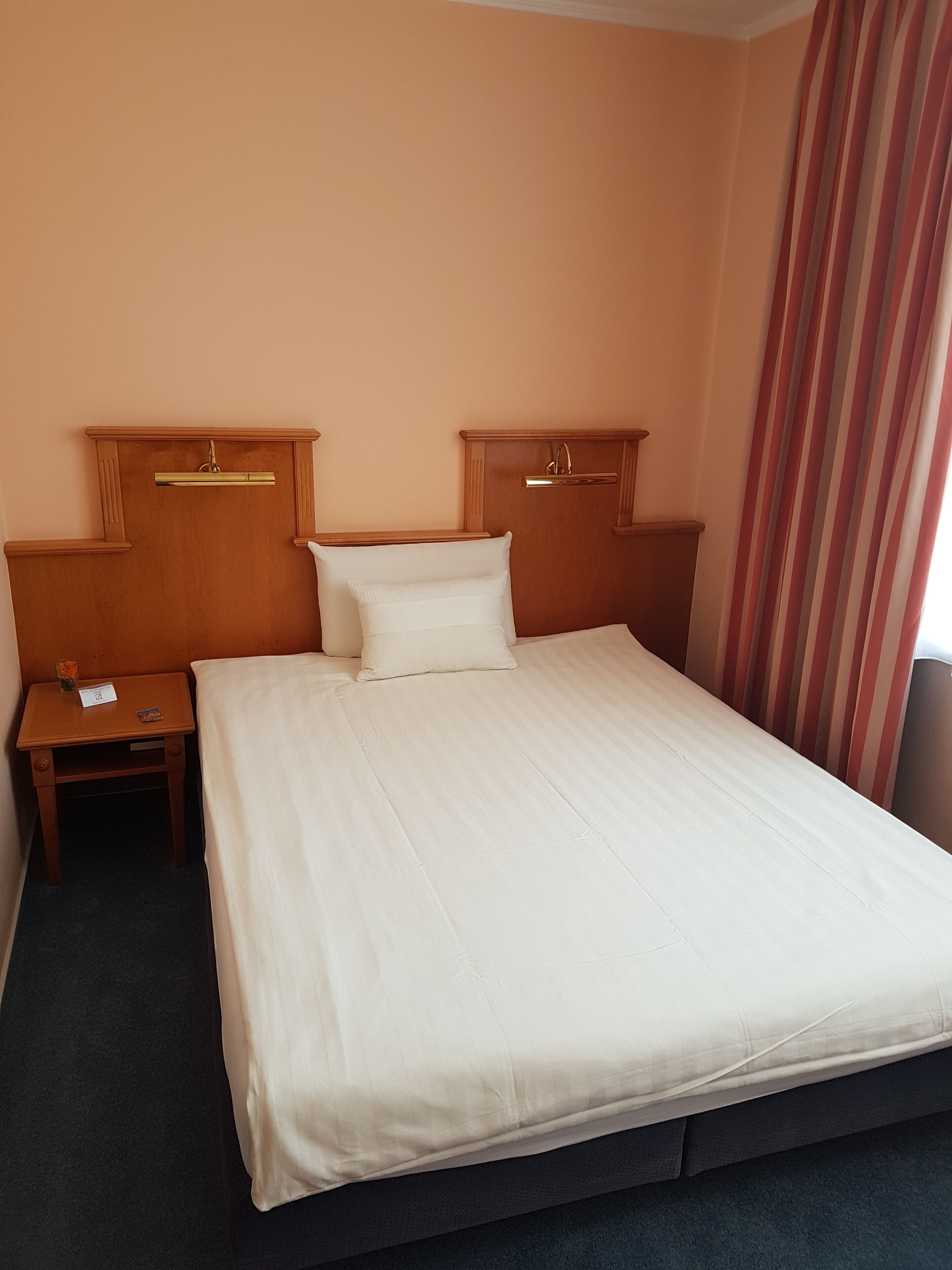 A hotel room with a large bed, wooden headboard, a nightstand, and a striped curtain.
