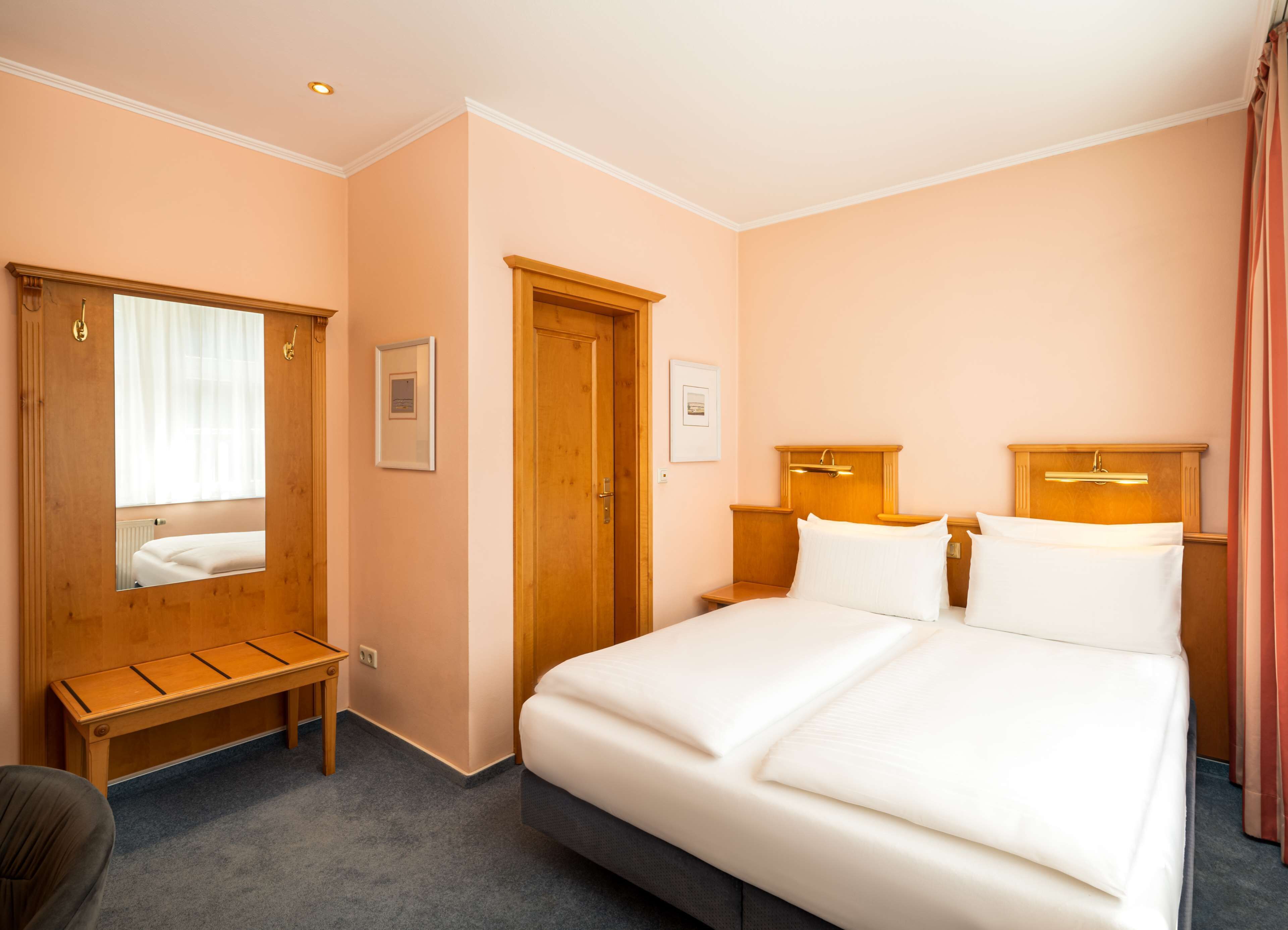 A hotel room with a double bed, a wooden bench, a wooden wardrobe, and a mirror.
