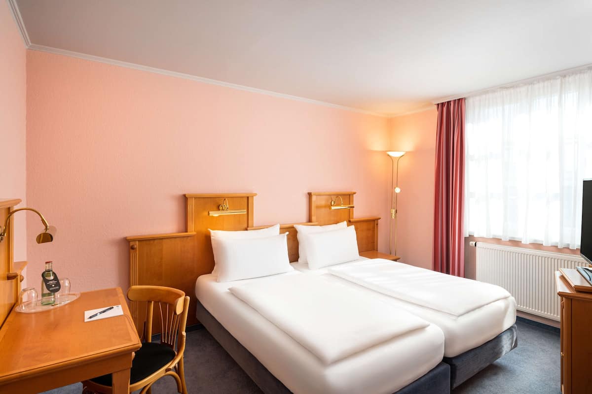 A hotel room with two beds, a desk with a chair, a lamp, a bottle of water, and a TV.