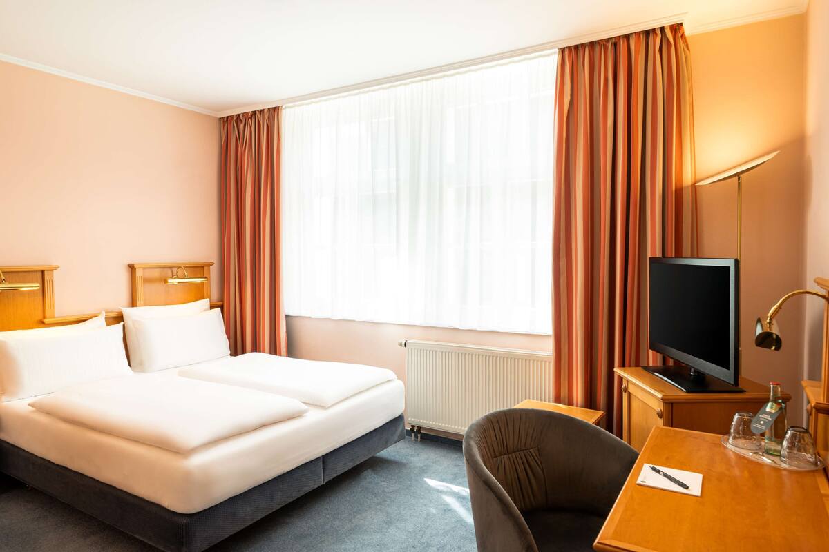 A hotel room with a double bed, a desk with a television, a chair, and a window with curtains.