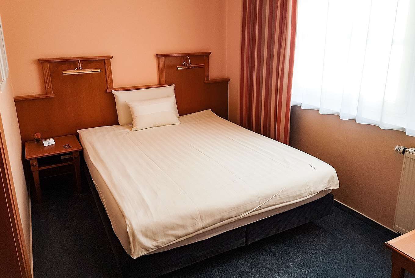 Habitación de hotel con una cama individual, cabecero de madera, mesitas de noche y una ventana con cortinas.