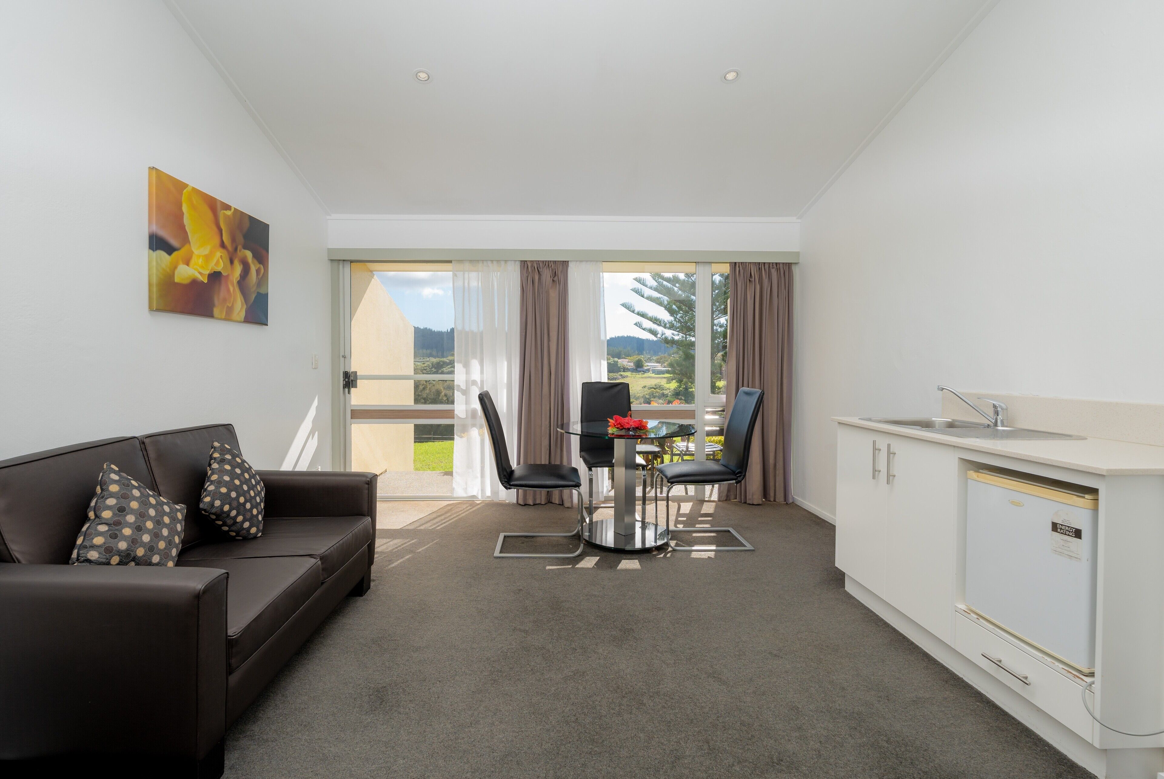 A modern hotel room with a brown leather sofa, a glass dining table with two chairs, a small kitchen area, and a large sliding glass door leading to a balcony.