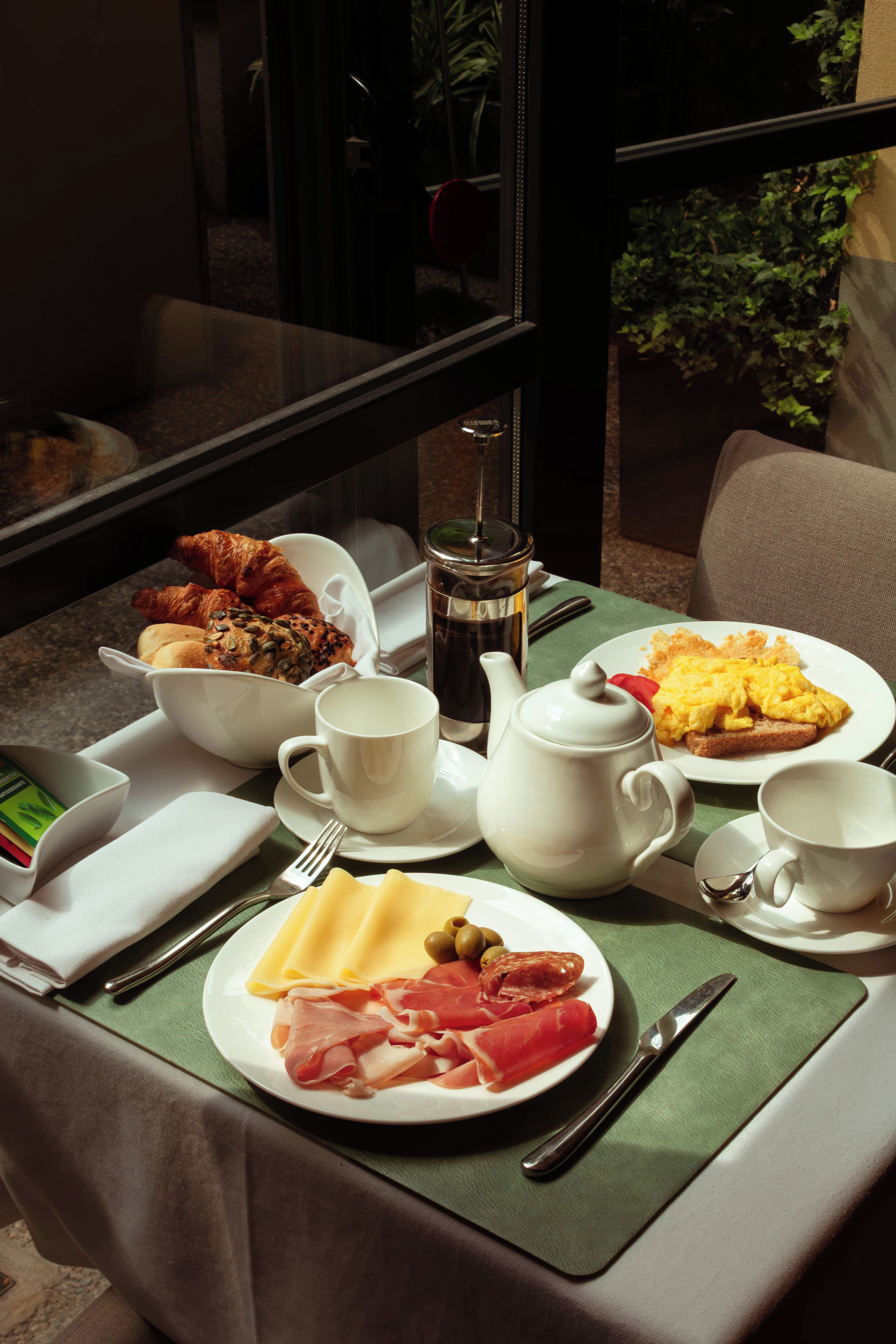 Una mesa de desayuno con platos de comida, tazas, una tetera y cubiertos.