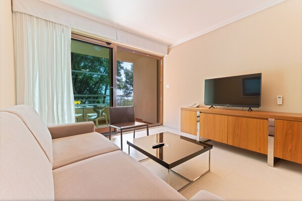 A modern living room with a sofa, a glass coffee table, a TV on a wooden cabinet, and a sliding glass door leading to a balcony. - The Residence (Podstrana)