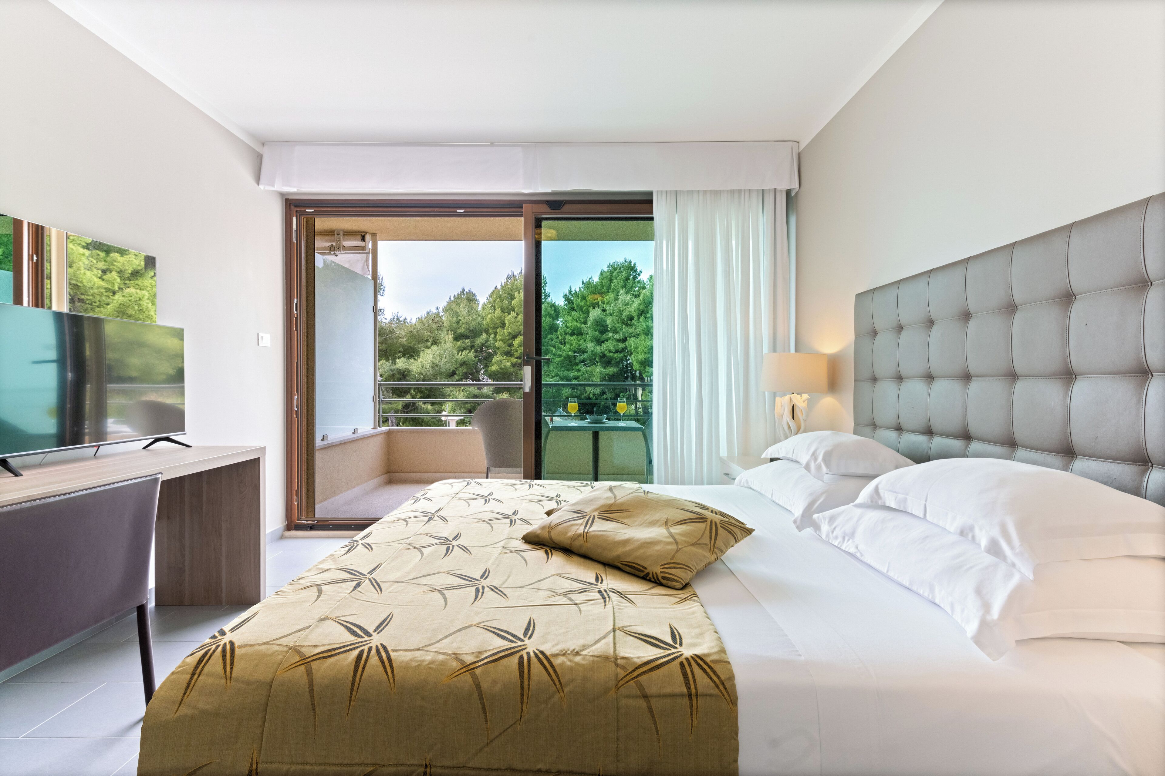 A hotel room with a bed, a desk, a television, and a balcony with a view of trees.