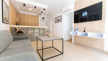 A modern reception area with a grey sofa, a glass-top table, wooden cabinets, and a mounted TV displaying the 'AURIANE' logo.