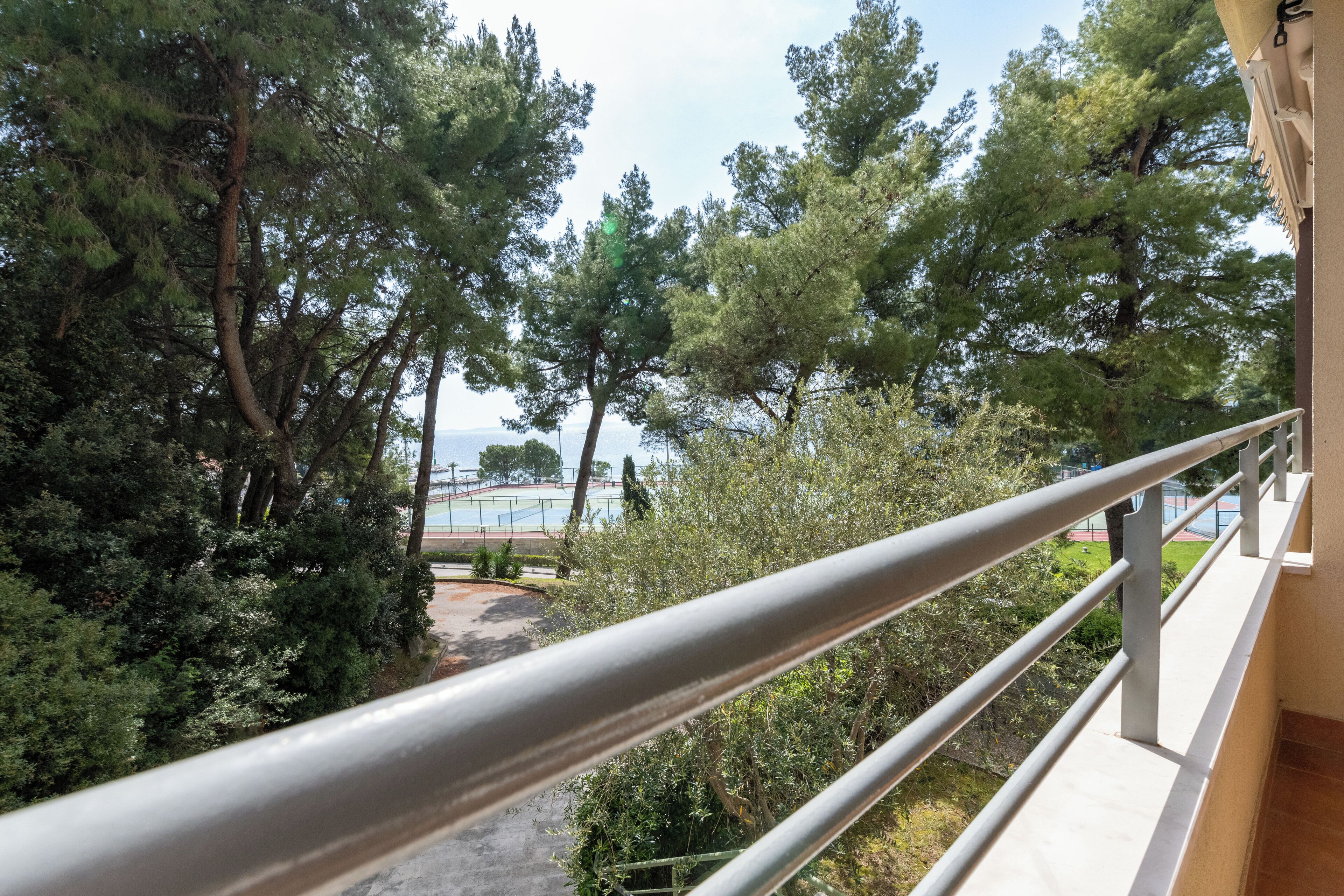 A balcony with a metal railing overlooking a garden with tall trees and a tennis court in the background.