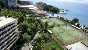 Ein Tennisplatz, umgeben von Grünflächen, neben einem Strand und einem Parkplatz.