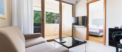A modern living room with a sofa, a glass coffee table, and a TV. There is a balcony with a view of trees and a chair outside.
