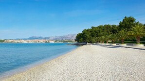 Ein Kiesstrand mit klarem, blauem Meer, einer Reihe von Palmen und einer in der Ferne liegenden Stadt mit Bergen.