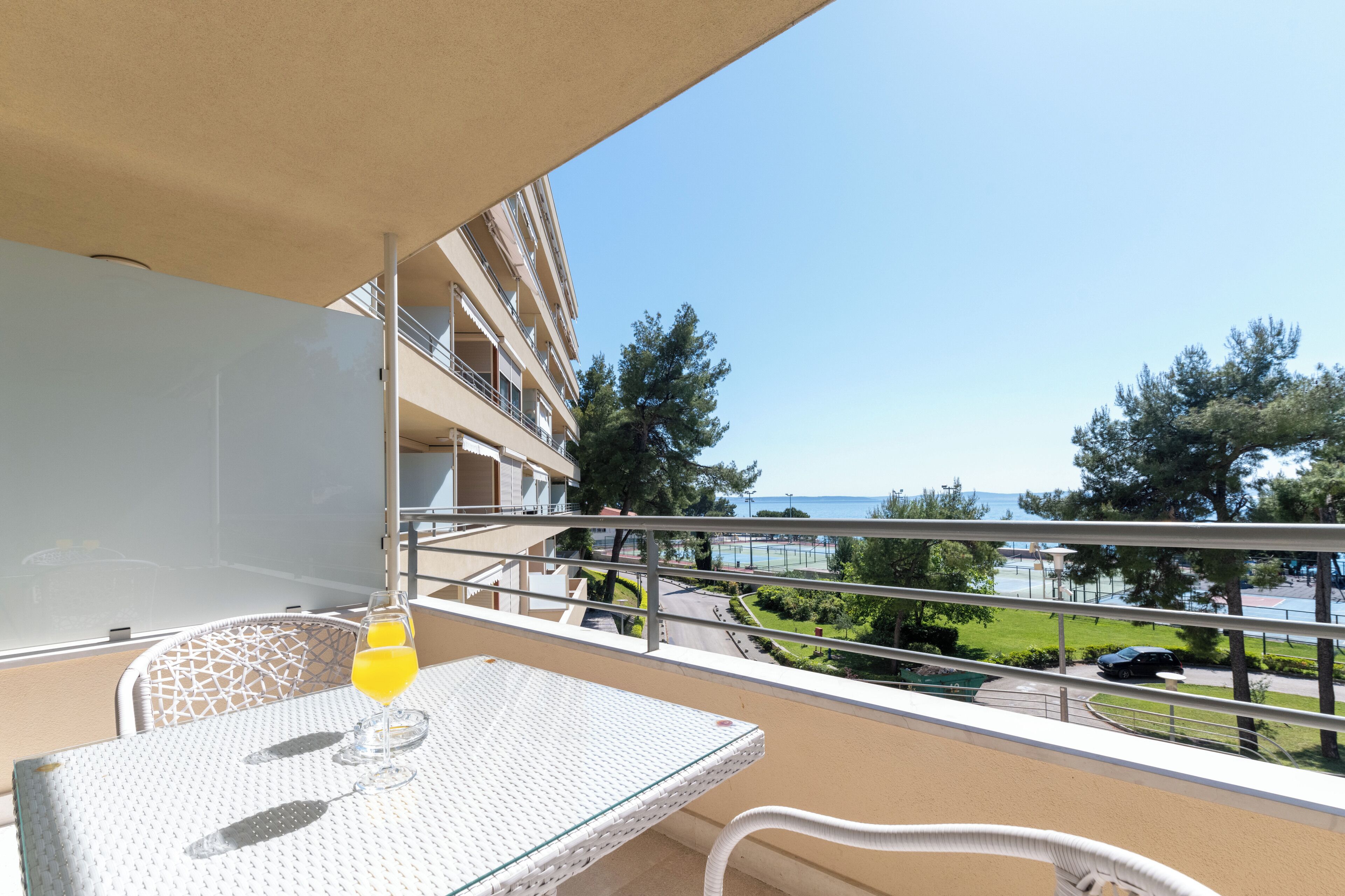 A balcony with a white wicker table, a glass of orange juice, and a view of the sea and a parking area.
