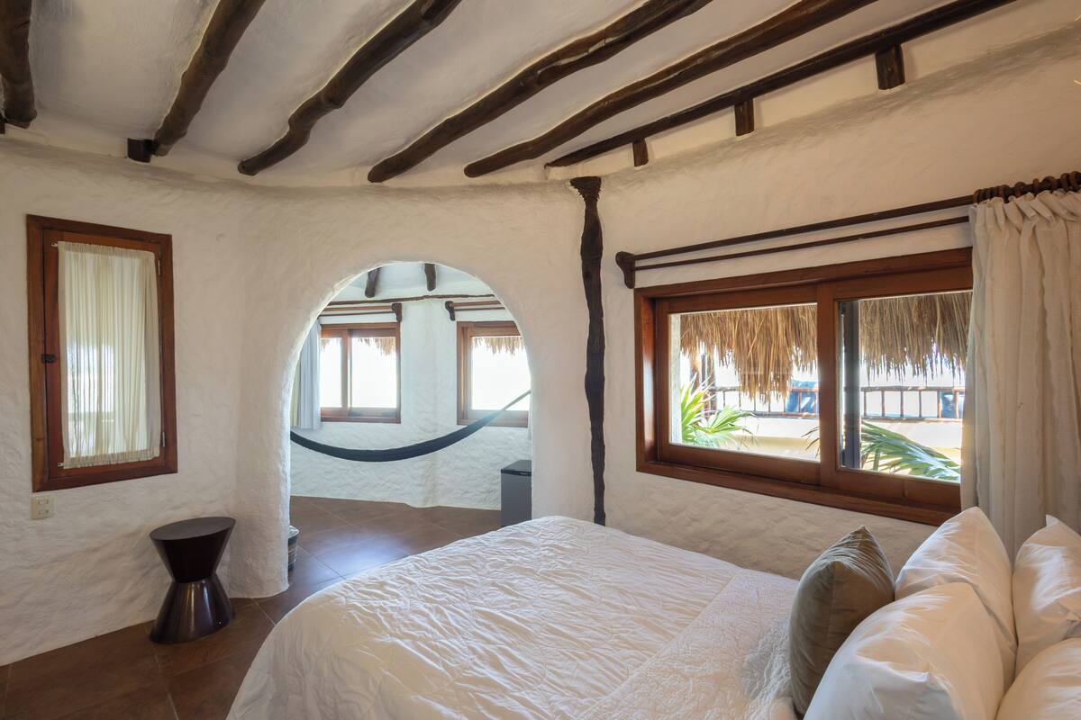 A bedroom with a bed, a window with curtains, a hammock, and a view of the beach.
